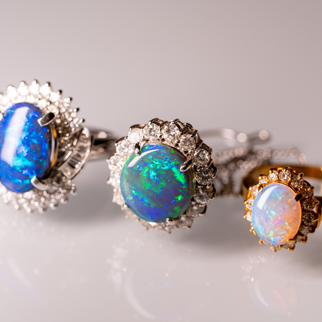 Drei Opalringe mit Diamanten, zwei aus Weißgold mit blauen und grünen Opalen, einer aus Gold mit einem orangefarbenen Opal, auf einem neutralen Hintergrund arrangiert.