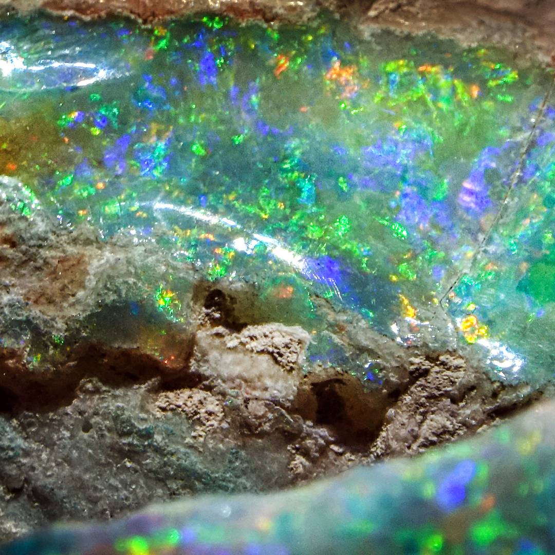 Detailaufnahme eines rohen Opals im Muttergestein, mit leuchtenden Farbspielen in Grün, Blau und Orange, die die natürliche Entstehung des Edelsteins fasziniert.