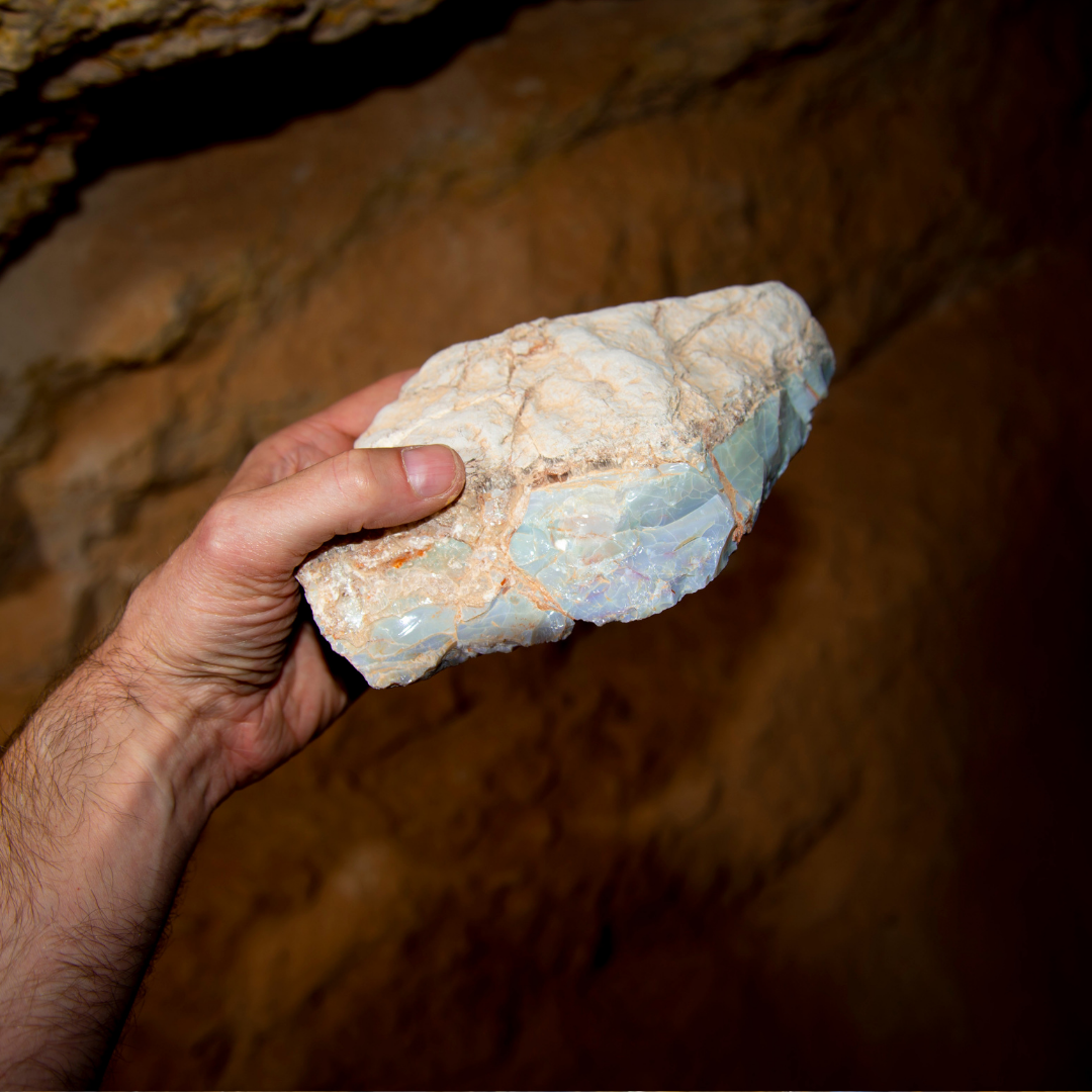 Hand, die ein großes Stück rohen Opal mit sichtbaren Adern in einem Minenschacht hält, brauner Hintergrund aus Erdschichten