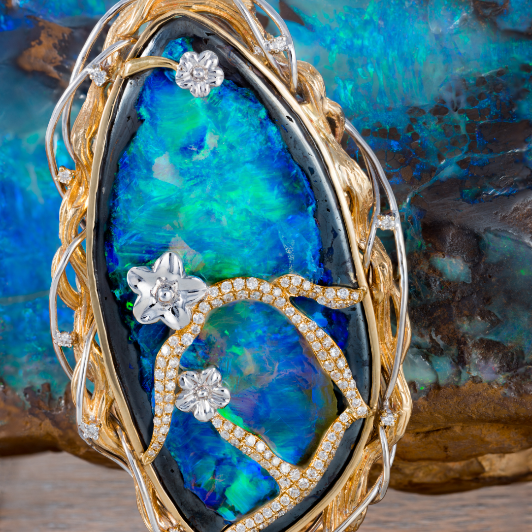 Hochwertiger Schmuckanhänger mit einem großen, intensiv schillernden Opal in Blau- und Grüntönen, verziert mit goldenen und silbernen Elementen sowie Diamanten, die Blumenmuster bilden, vor dem Hintergrund eines ungeschliffenen Rohopals.