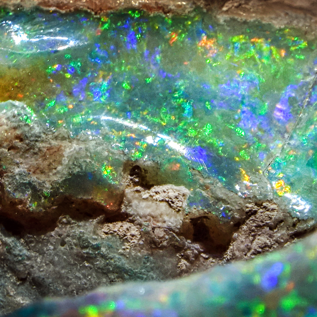 Nahaufnahme eines Opals mit lebhaften, schimmernden Farben in Grün-, Blau- und Orangetönen, eingebettet in Muttergestein