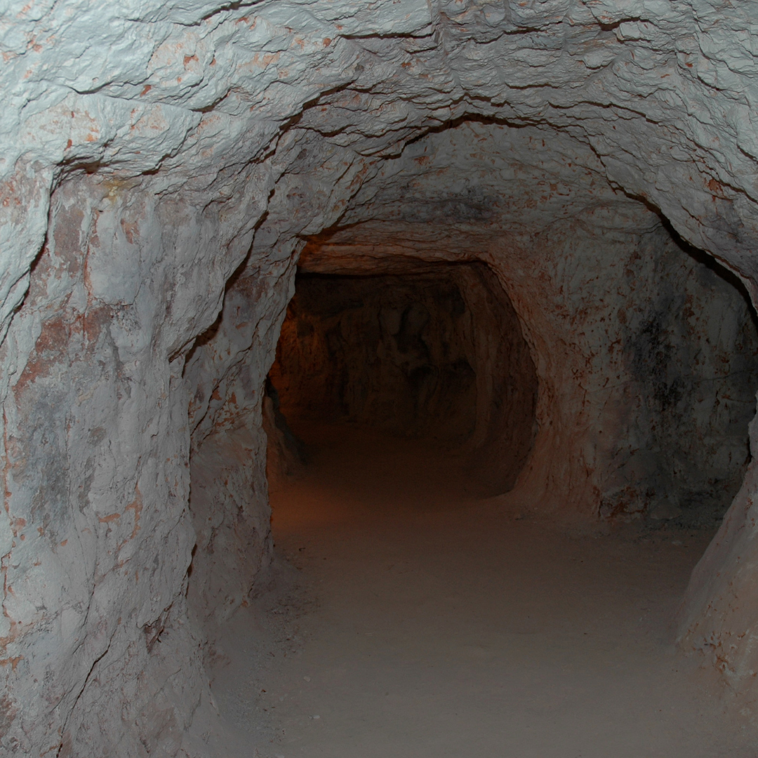 Unterirdischer Minenschacht von Opalen mit rauen, ungleichmäßigen Wänden aus hellem Gestein, schwach beleuchtet, mit einem dunkleren Tunnelverlauf im Hintergrund.