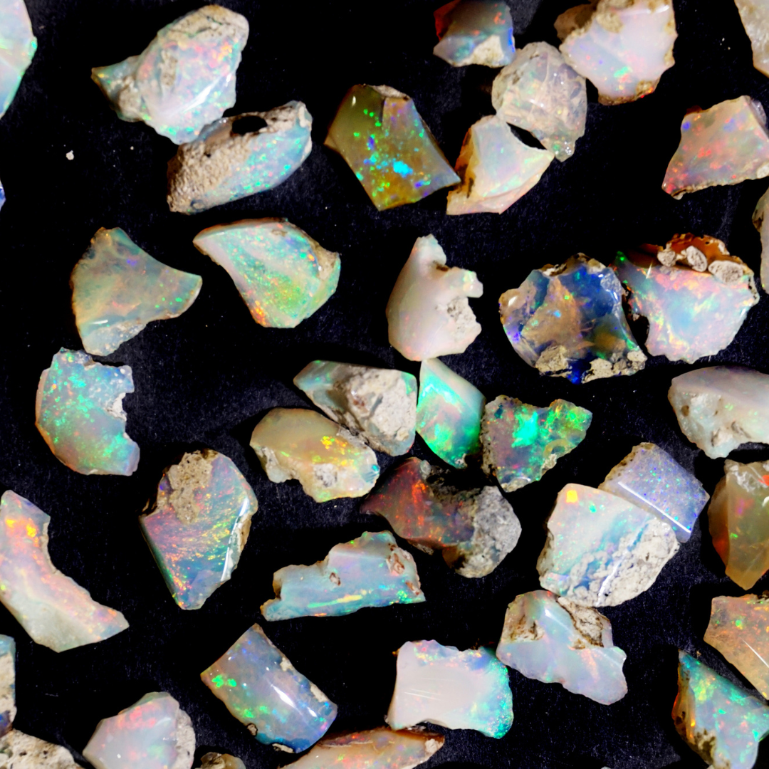 Verschiedene rohe Opalstücke auf schwarzem Hintergrund, die durch ihre bunten Farbreflexe in Blau, Grün und Rot auffallen. Die ungeschliffenen Steine zeigen die natürliche Vielfalt und Schönheit des Edelsteins in seiner ursprünglichen Form.
