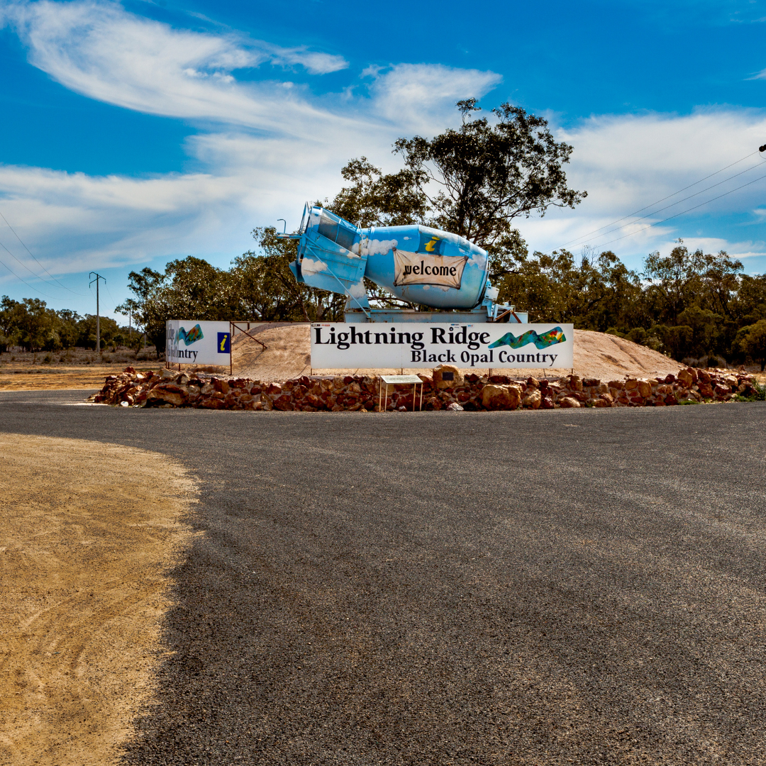Willkommensschild für Lightning Ridge, Black Opal Country, auf einem Kreisverkehr mit einer bemalten Betonmischmaschine als Skulptur, umgeben von Bäumen unter einem blauen Himmel.